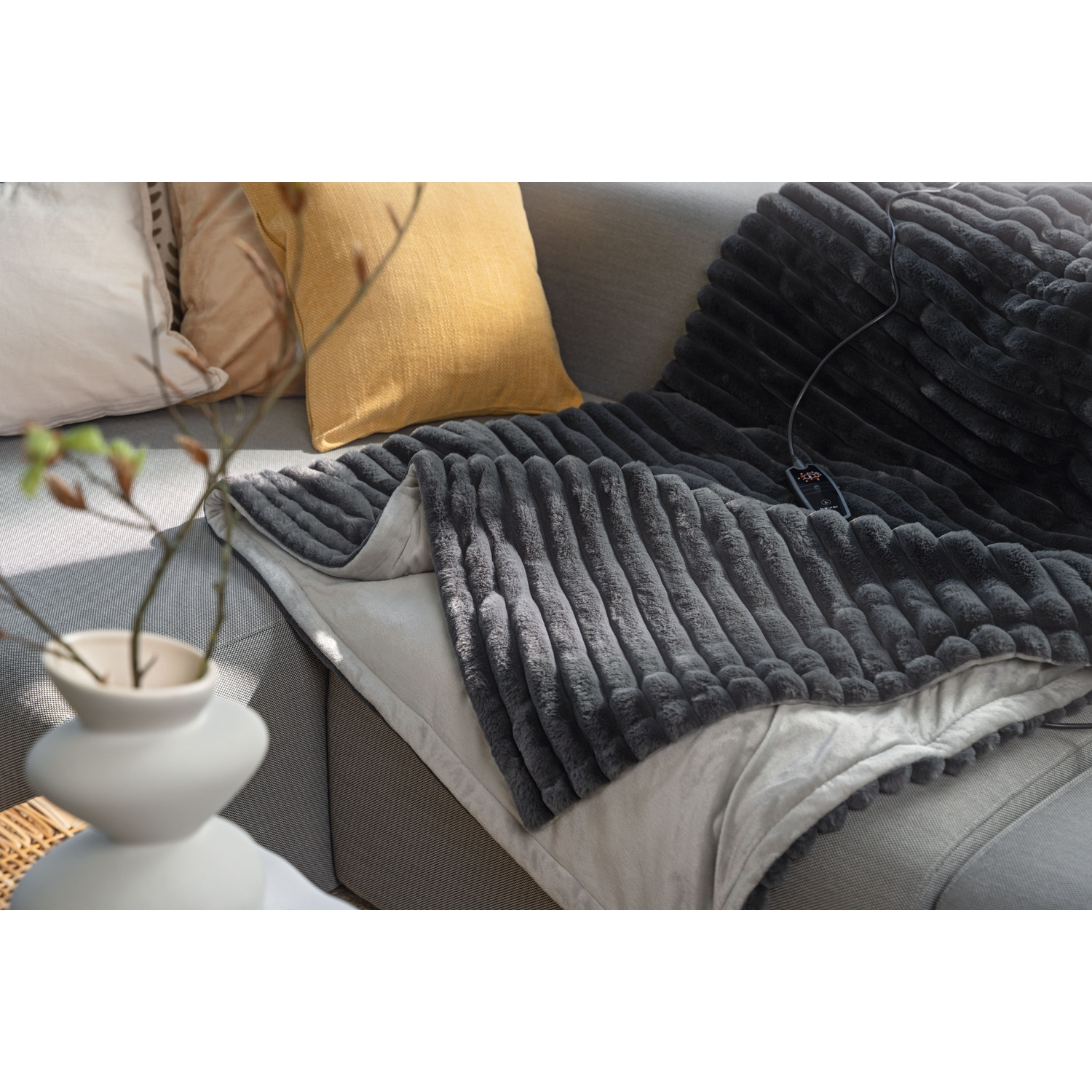 A gray blanket with a controller is on a gray sofa. Yellow and white pillows are in the background, with a vase of branches.