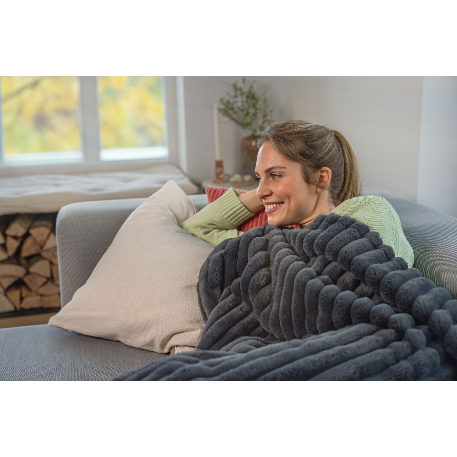 A woman smiles while relaxing on a gray sofa, covered by a gray blanket. A window and pillows are in the background.