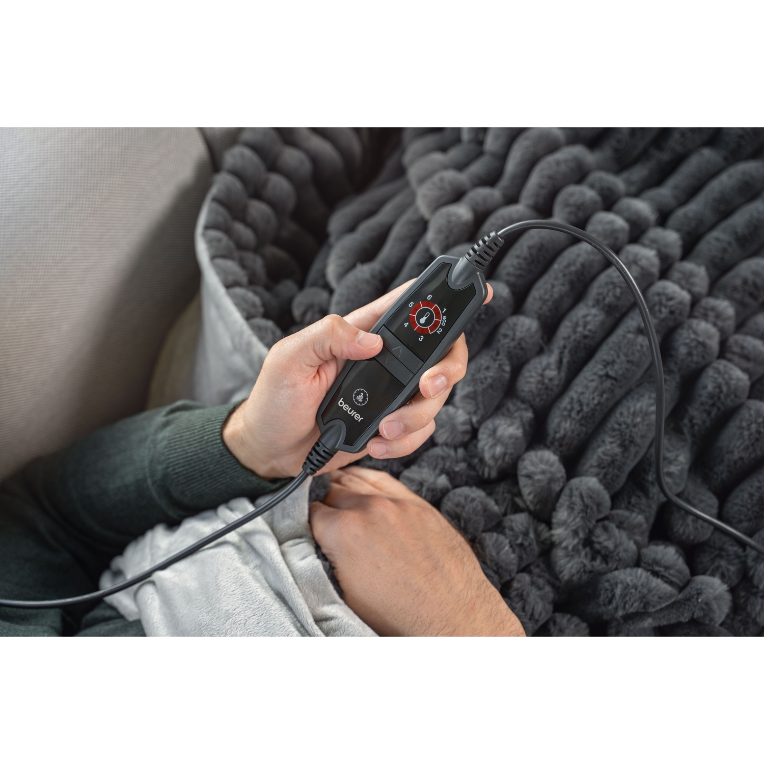 A person holds a black controller with a dial. It's connected to a gray plush blanket. The person is wearing a green sweater.