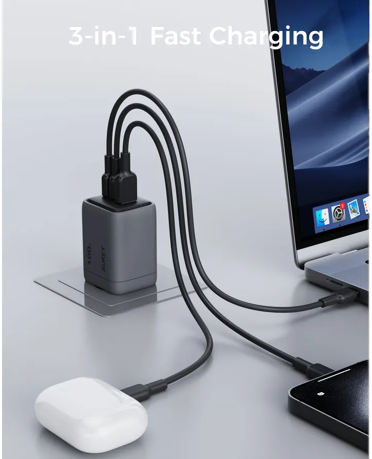 Un caricatore rapido 3-in-1 è collegato a un laptop, un telefono e auricolari. Il caricatore è grigio e lo sfondo è una scrivania grigio chiaro.