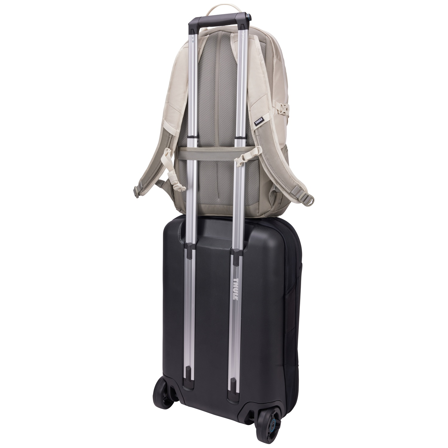 Een beige rugzak rust op een zwarte rolkoffer. Beide staan tegen een witte achtergrond.