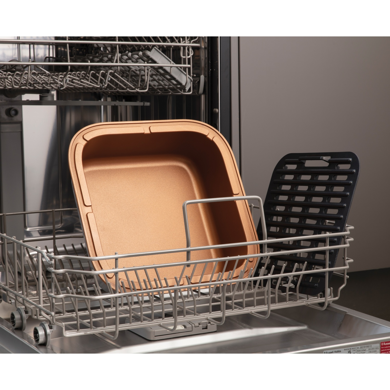 Lavastoviglie con tegame in rame e griglia nera all'interno. La macchina è aperta, con interno grigio. Il tegame è quadrato, la griglia rettangolare.