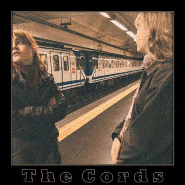 Zwei Frauen auf einem U-Bahnhof, Zug im Hintergrund. Eine Frau blickt nach oben, die andere nach vorne. Titel unten: The Cords.
