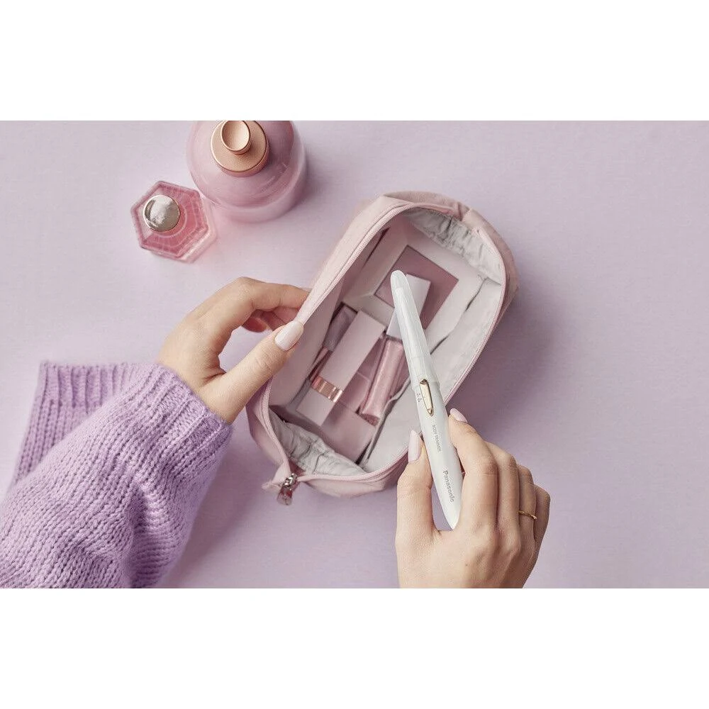 Manos sosteniendo una herramienta de belleza Panasonic blanca. Una bolsa rosa está abierta, revelando artículos de maquillaje. También se ven frascos de perfume y un suéter morado.