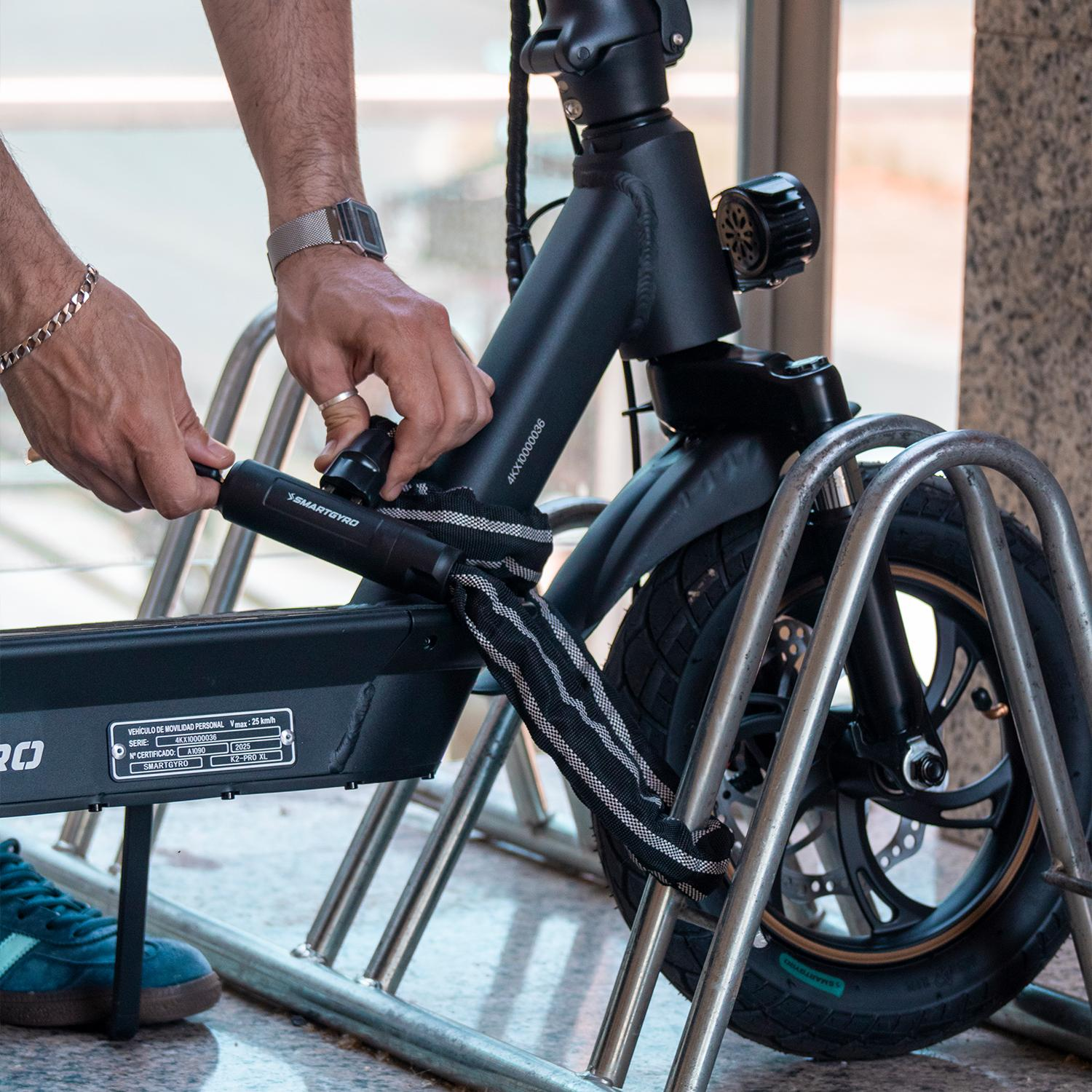 Una persona asegura un patinete eléctrico a un soporte metálico. El patinete es negro y se ven las manos de la persona. La escena es al aire libre.