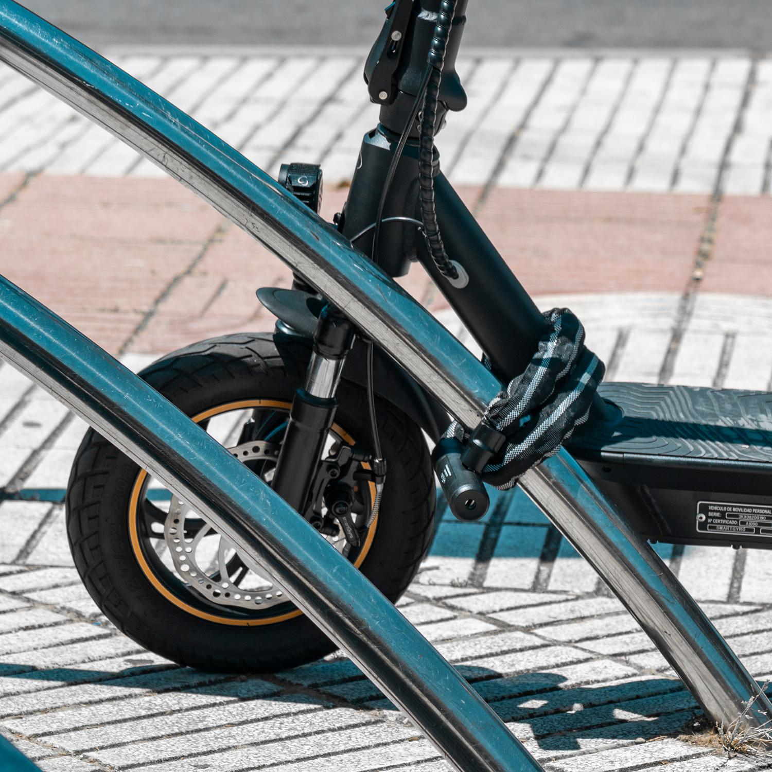 Un patinete eléctrico asegurado a una barra de metal. El patinete es negro con detalles dorados y un candado blanco y negro.