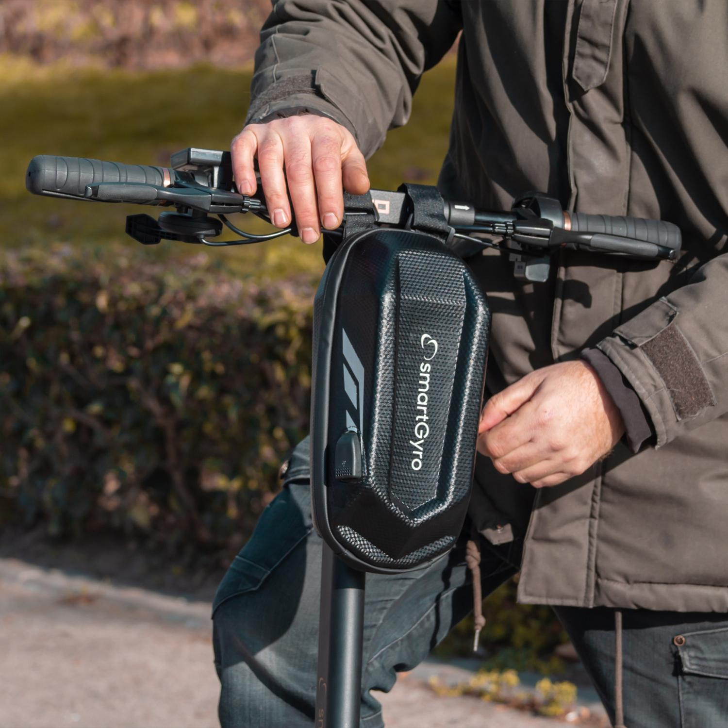 Una persona sostiene un patinete eléctrico. El patinete tiene una bolsa negra que dice SmartGyro. La persona lleva chaqueta y vaqueros.