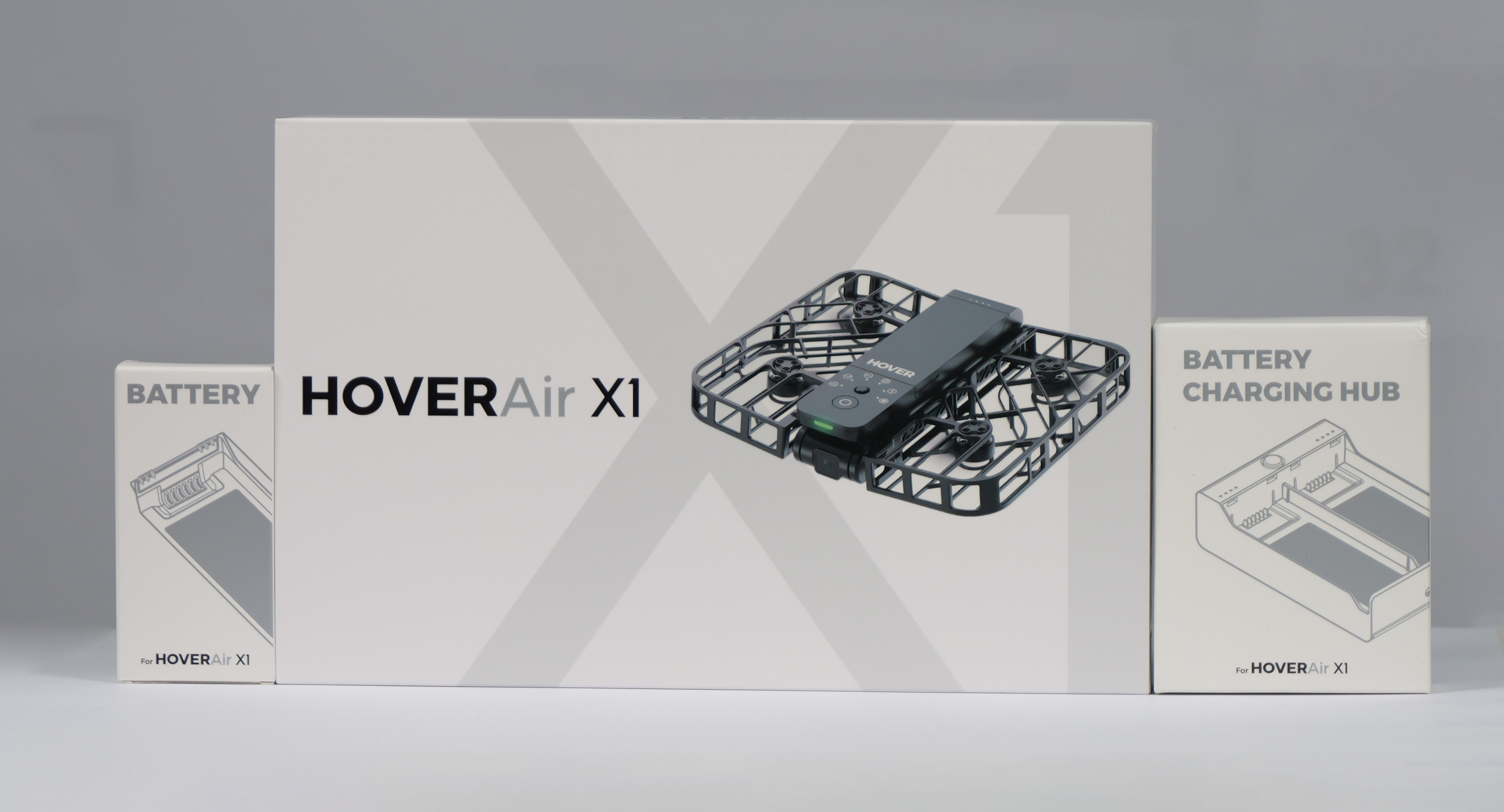 La imagen muestra un dron HoverAir X1 con cajas de batería y centro de carga. El dron es negro y las cajas son blancas.