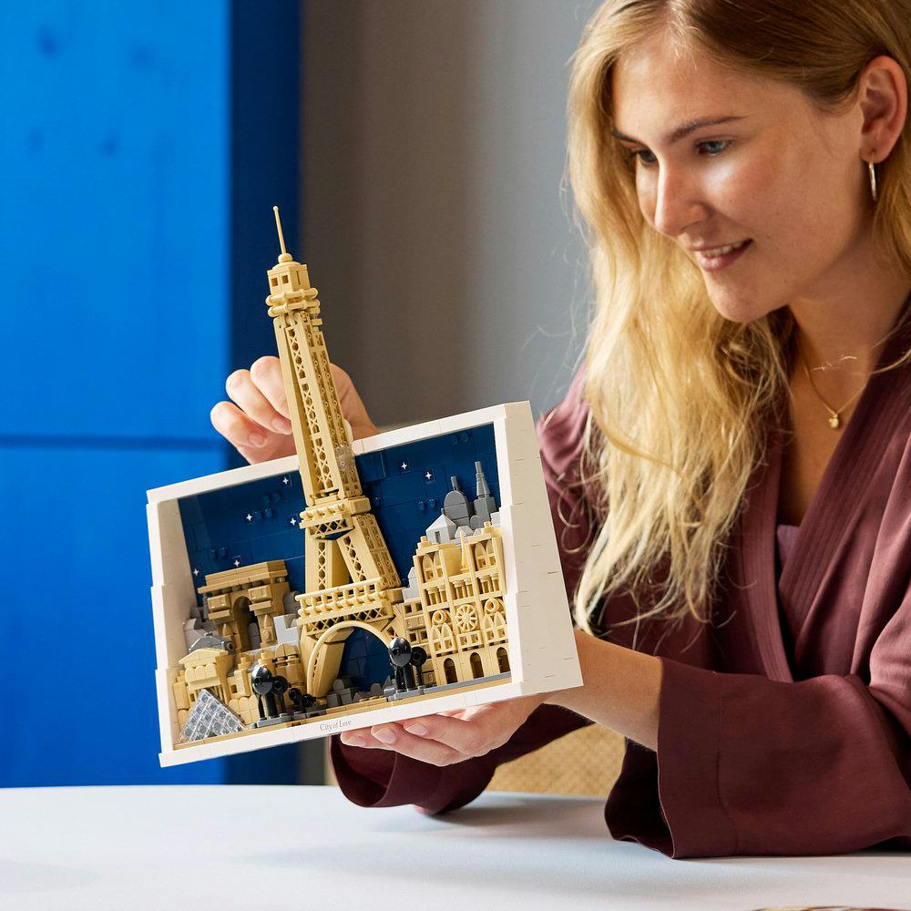 Eine Frau hält ein Lego-Modell von Paris. Das Modell zeigt den Eiffelturm, den Arc de Triomphe und andere Gebäude vor blauem Hintergrund.