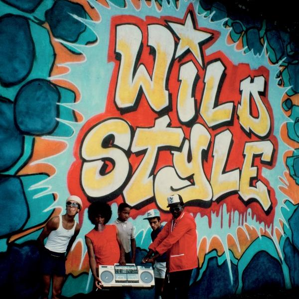 Gruppe von Leuten an einer Graffiti-Wand. An der Wand steht "Wild Style". Ein Mann hält eine Boombox. Die Leute tragen Hüte und Sonnenbrillen.