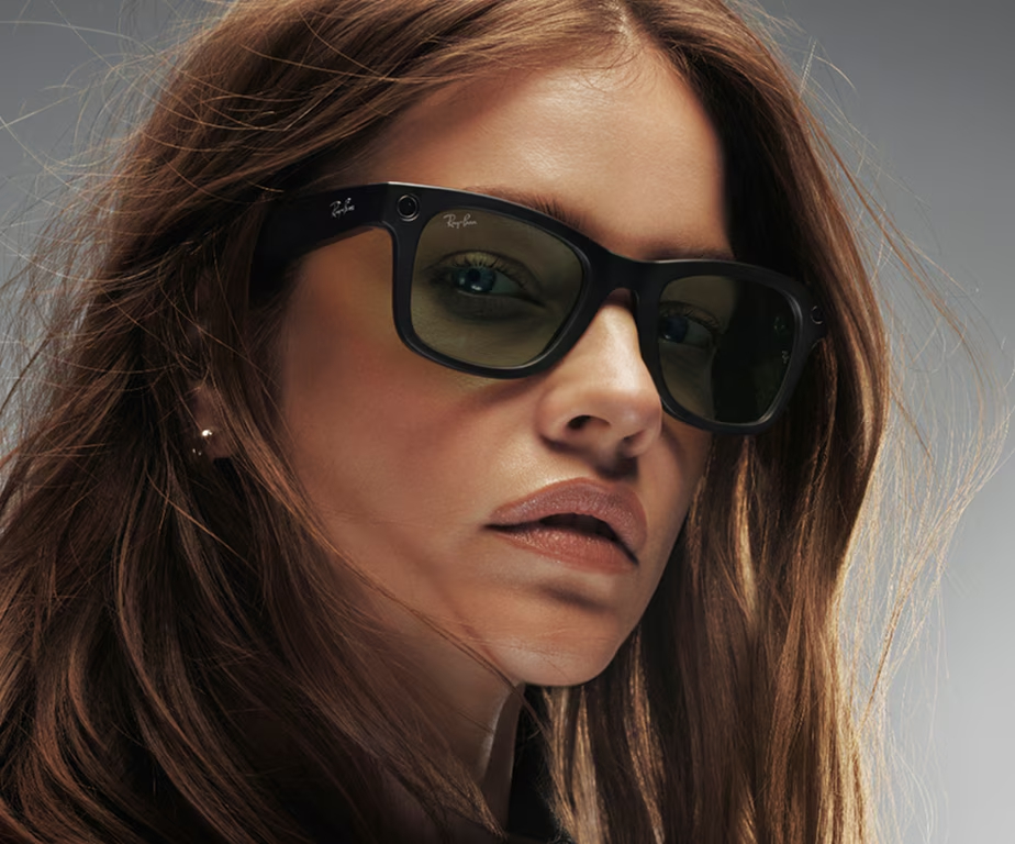 Kobieta w czarnych okularach przeciwsłonecznych Ray-Ban, patrząca w kamerę. Ma brązowe włosy i jasne tło. Okulary są na niej.