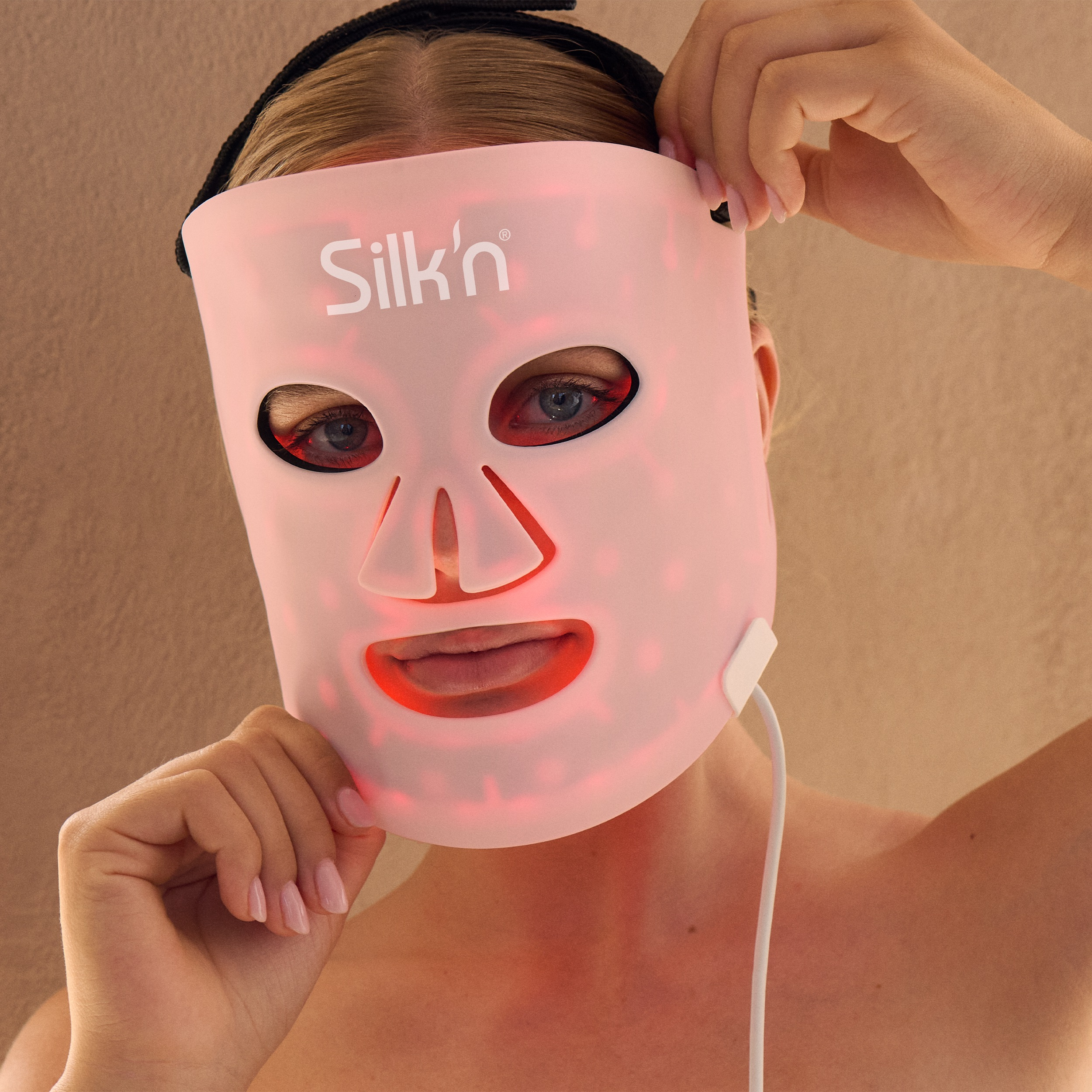 Kobieta w różowej masce LED z logo 'Silk'n'. Czerwone światła świecą. Dłonie trzymają maskę.