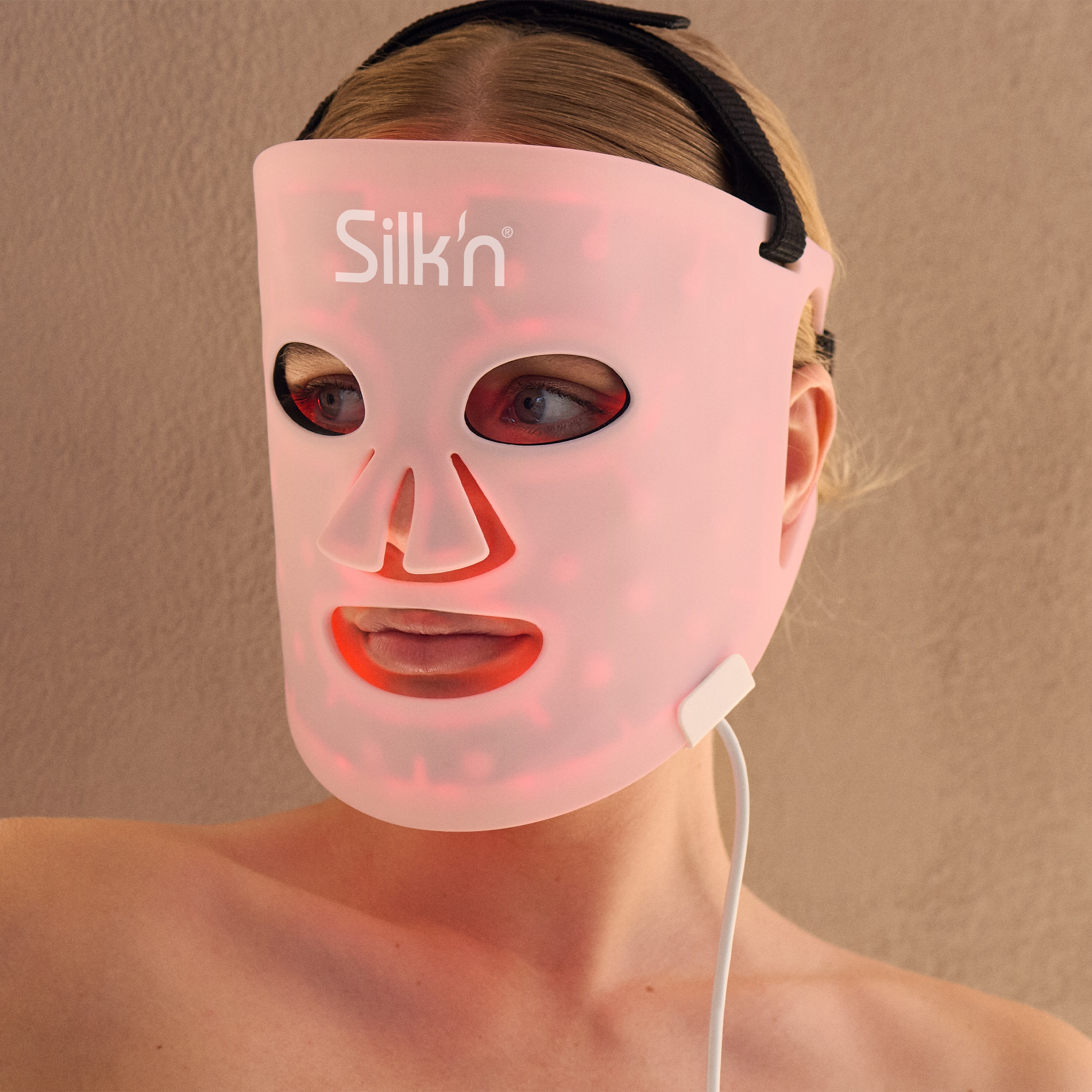 Kobieta nosi różową maskę do twarzy LED Silk'n. Widoczne są blond włosy. Biały kabel zwisa z maski.