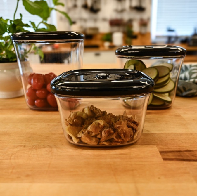 Trzy szklane pojemniki z czarnymi pokrywkami na drewnianej powierzchni. Zawierają pomidory, plasterki cukinii i gotowane jedzenie.