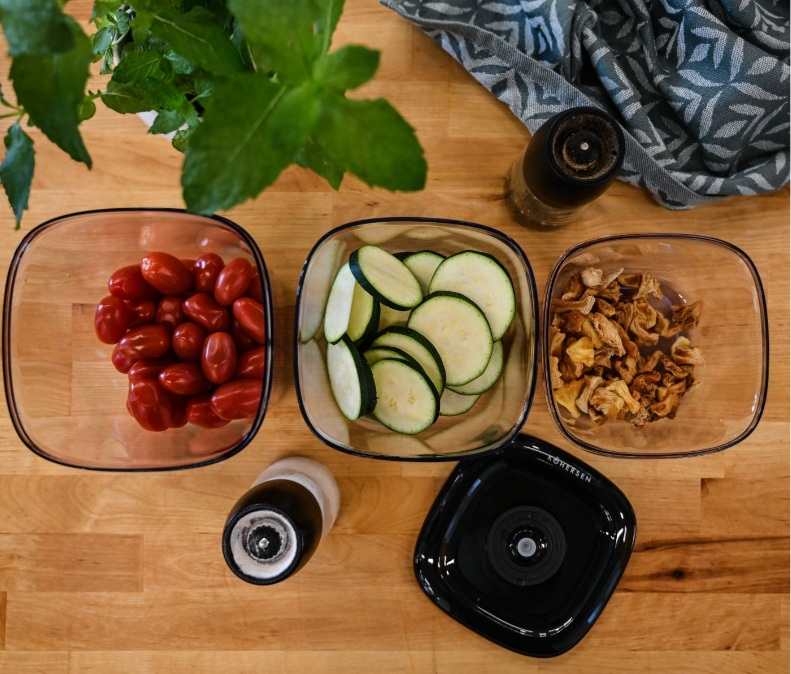 Widok z góry na warzywa w szklanych miskach: pomidory, cukinia i suszone owoce. Zioła i ręcznik w tle.