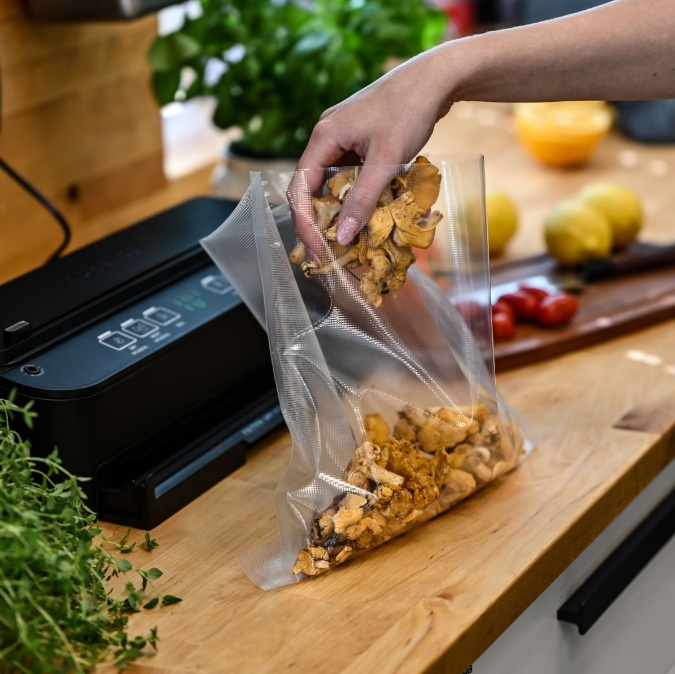 Ręka umieszcza kurki w przezroczystej plastikowej torbie. Czarna zgrzewarka próżniowa w tle na drewnianej powierzchni.