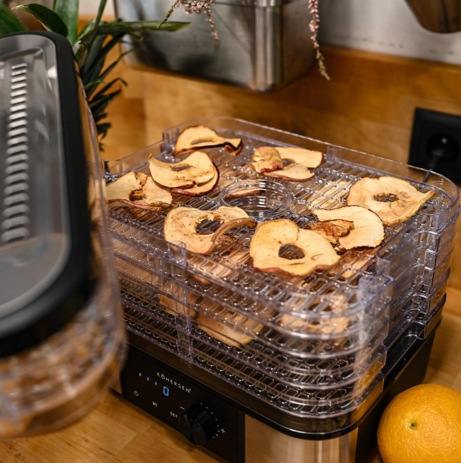 Suszarka do owoców z plasterkami jabłek. Czarna i przezroczysta maszyna. Pomarańcza jest w prawym rogu. Tło z rośliną i elementami kuchennymi.