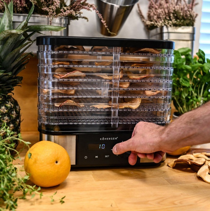 Suszarka do żywności z suszonymi owocami w środku, ręka regulująca ustawienia. Na stole grejpfrut i ananas.