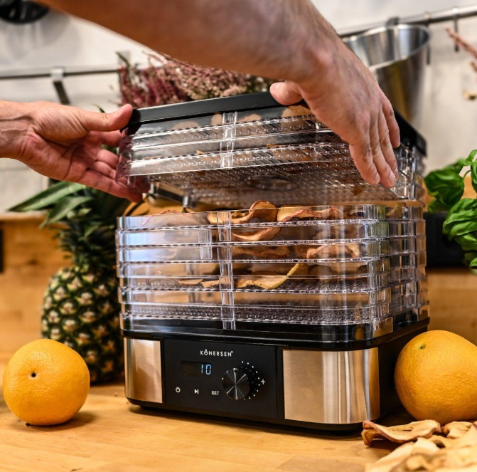 Osoba otwiera suszarkę do żywności. W środku znajdują się suszone plasterki jabłka. Jest ananas, dwie pomarańcze oraz czarno-srebrna maszyna.