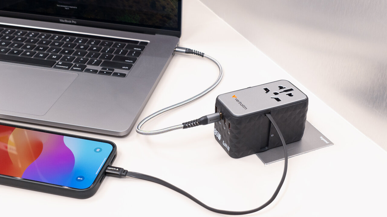 Beyaz bir yüzeyde şarj olan dizüstü bilgisayar, telefon ve güç adaptörü. Kablolar cihazları bağlar. Adaptör siyah ve gümüş rengindedir.