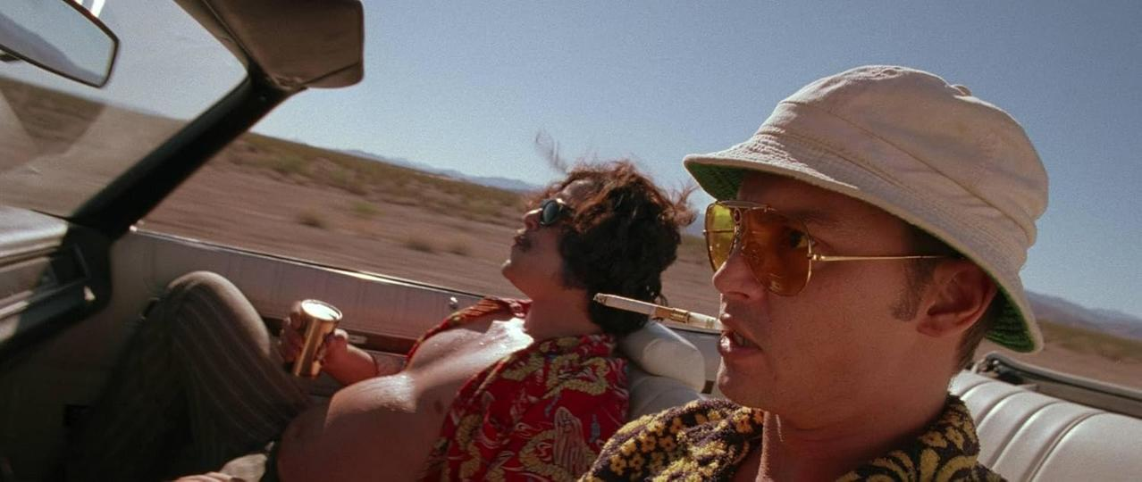 Zwei Männer in einem Cabrio, einer raucht, der andere hat ein Getränk. Wüstenhintergrund, sonniger Tag.