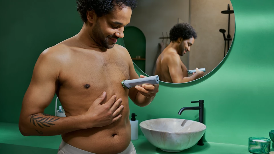 Ein Mann rasiert sich im Badezimmer die Brust. Er hält einen Trimmer und lächelt. Ein Spiegel spiegelt sein Bild wider. Grüne Wände und ein Waschbecken sind zu sehen.