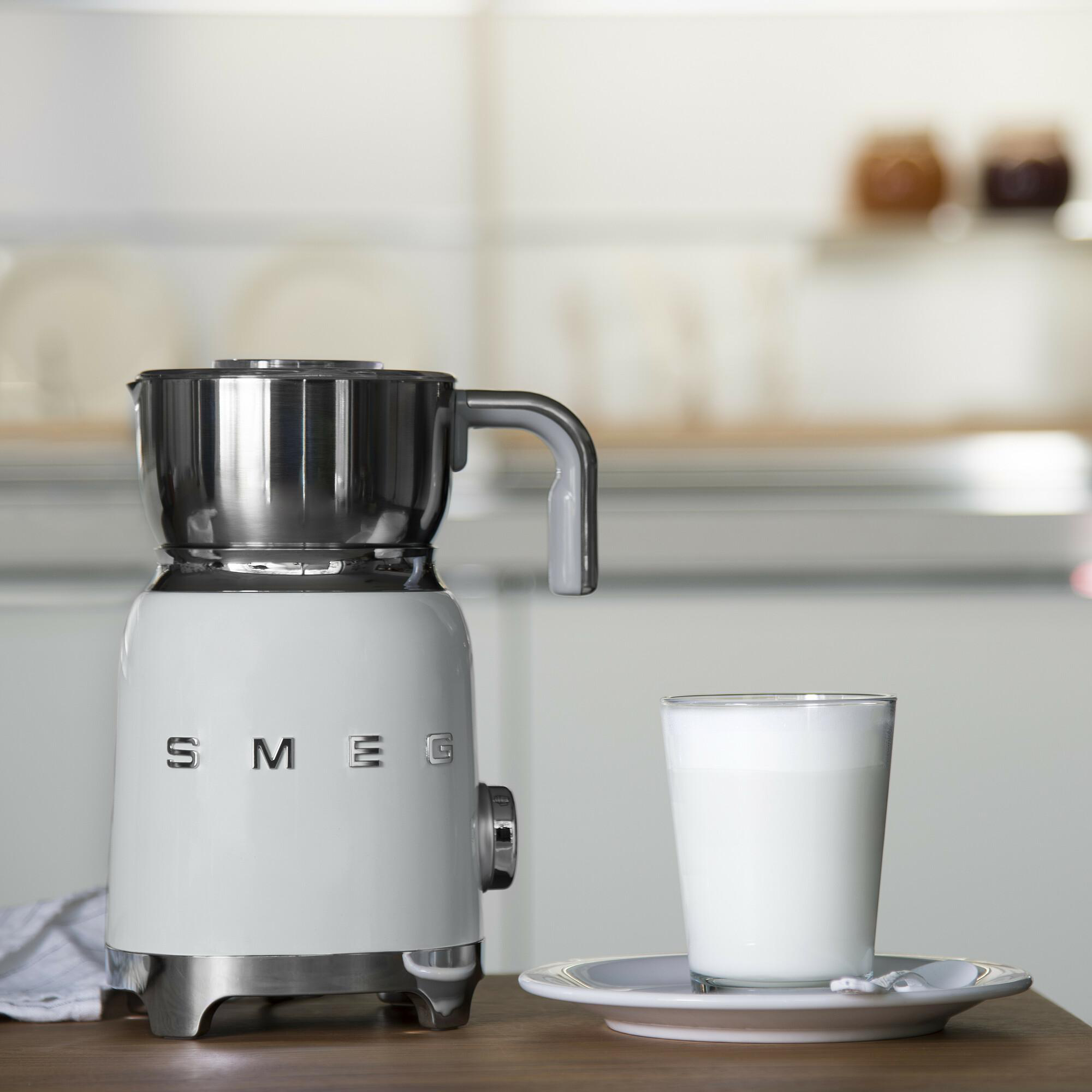 Ein weißer SMEG-Milchaufschäumer, ein Glas Milch auf einer Untertasse und eine Küchenumgebung im Hintergrund.