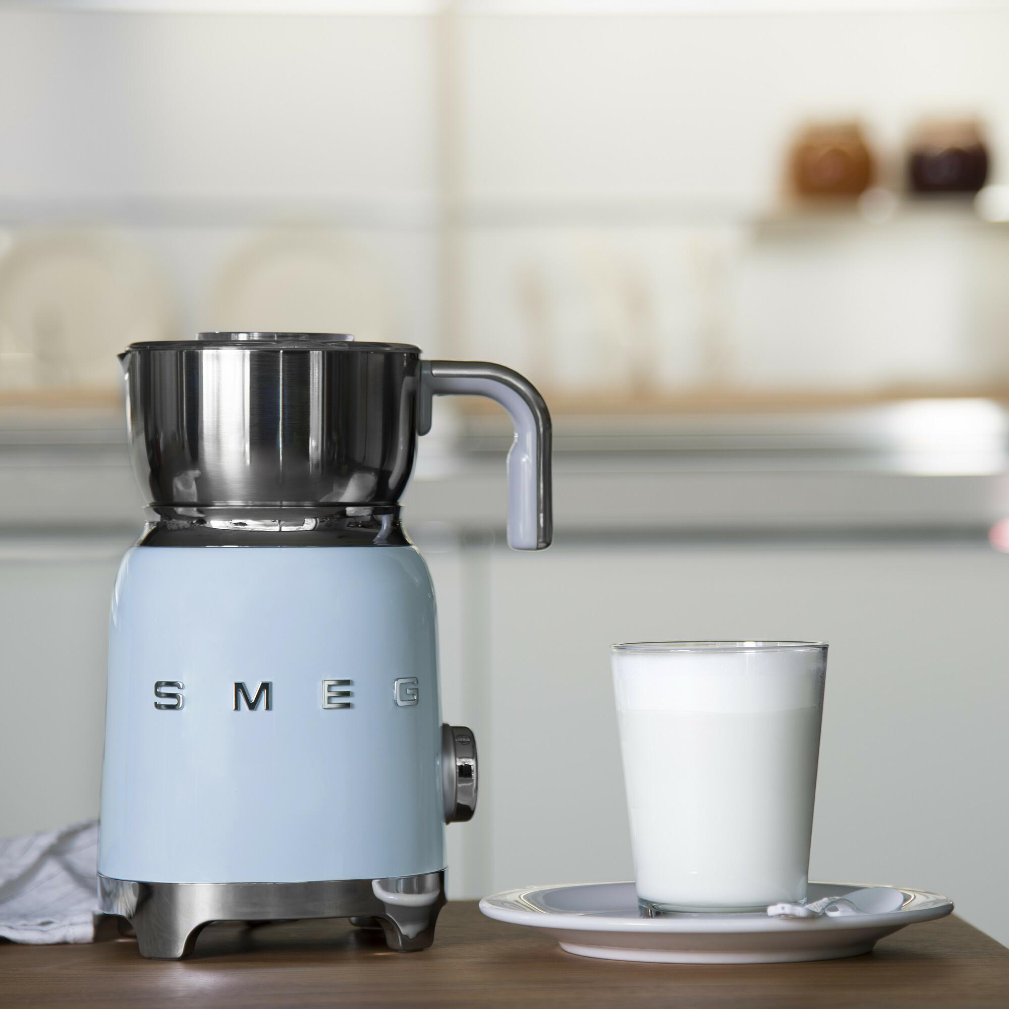 Ein hellblauer SMEG-Milchaufschäumer und ein Glas Milch auf einem Holztisch. Hintergrund zeigt eine Küche.