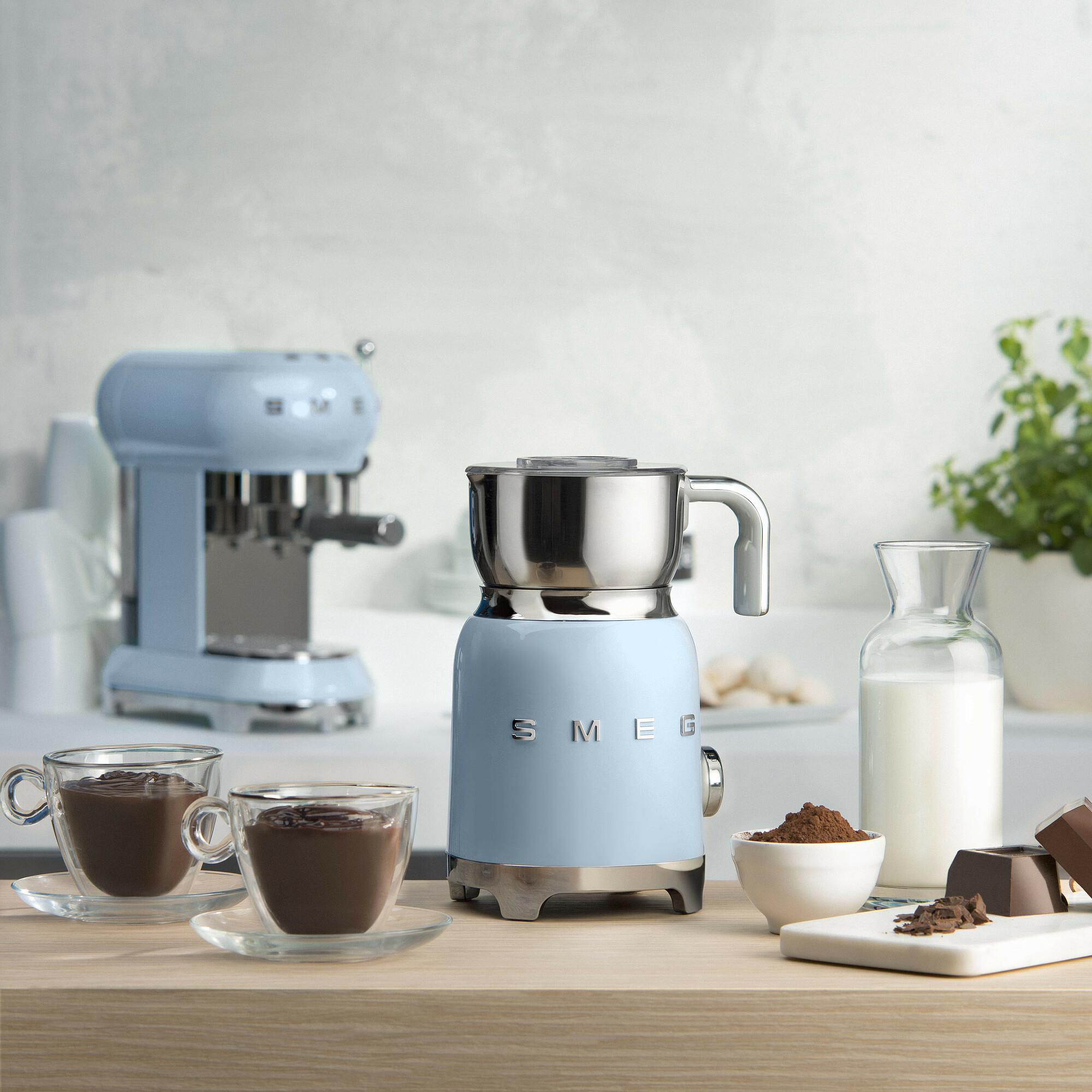 Ein hellblauer SMEG-Milchaufschäumer, Espressomaschine, Tassen mit heißer Schokolade, Milch und Schokoladenstücke auf Holz.