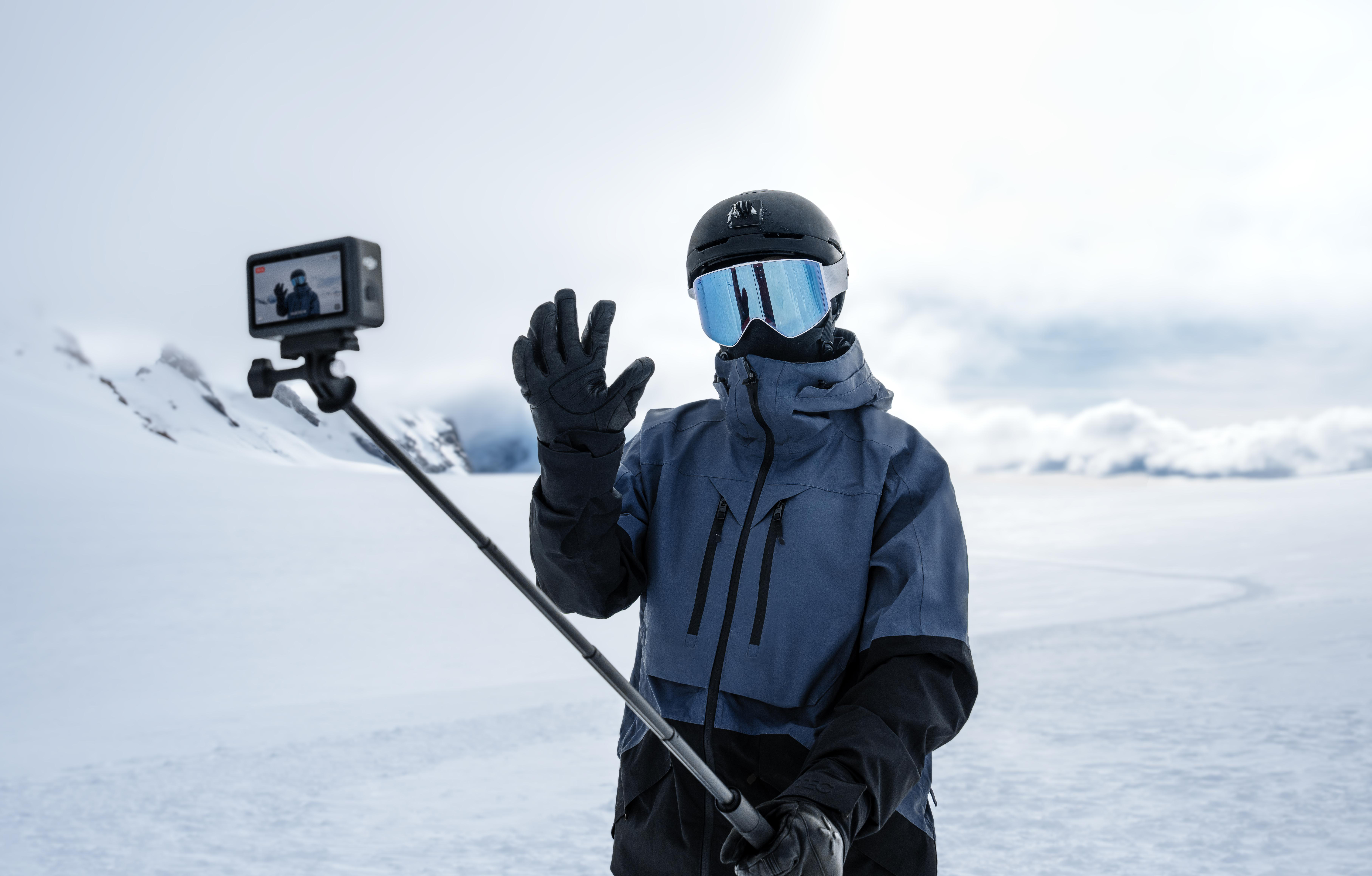 Skifahrer macht Selfie in schneebedeckten Bergen. Er winkt, trägt Helm, Brille und blaue Jacke. Kamera auf Selfie-Stick.