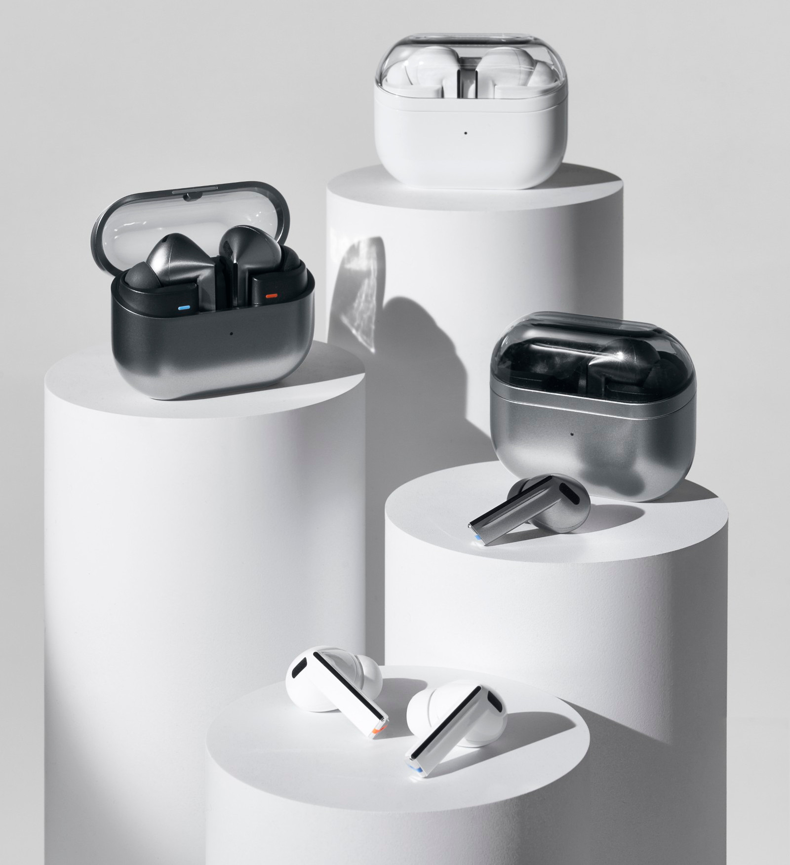 Słuchawki douszne w etui na cokołach. Etui są białe i srebrne, słuchawki białe i szare. Czysta, nowoczesna estetyka.