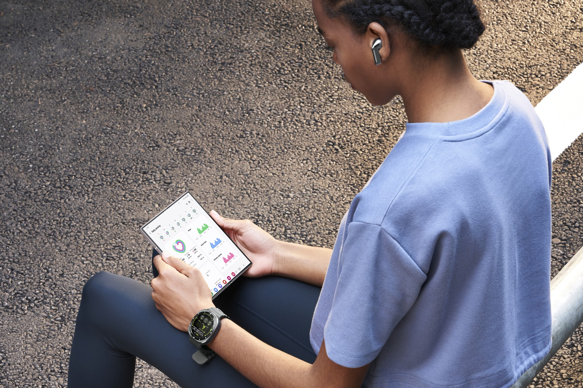 Osoba siedzi na zewnątrz i patrzy na tablet z danymi aktywności. Ma na sobie smartwatch, słuchawki i niebieską koszulę.