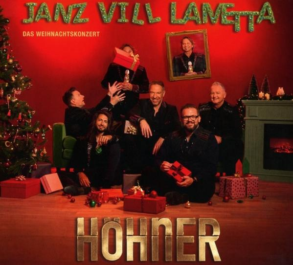 Eine Band posiert vor einer Weihnachtsdekoration. Sie sind von Geschenken und einem Weihnachtsbaum umgeben. Der Bandname ist 'Hohner'.