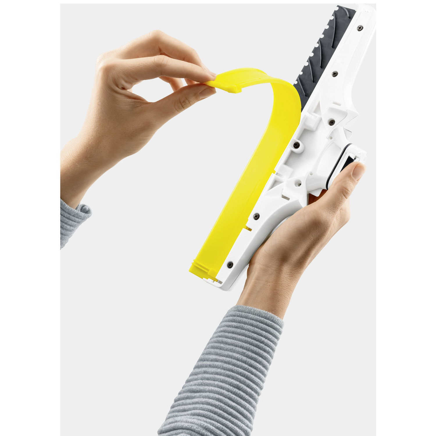 Hands hold a white and black cleaning tool with yellow detail. One hand peels a yellow strip from the tool.