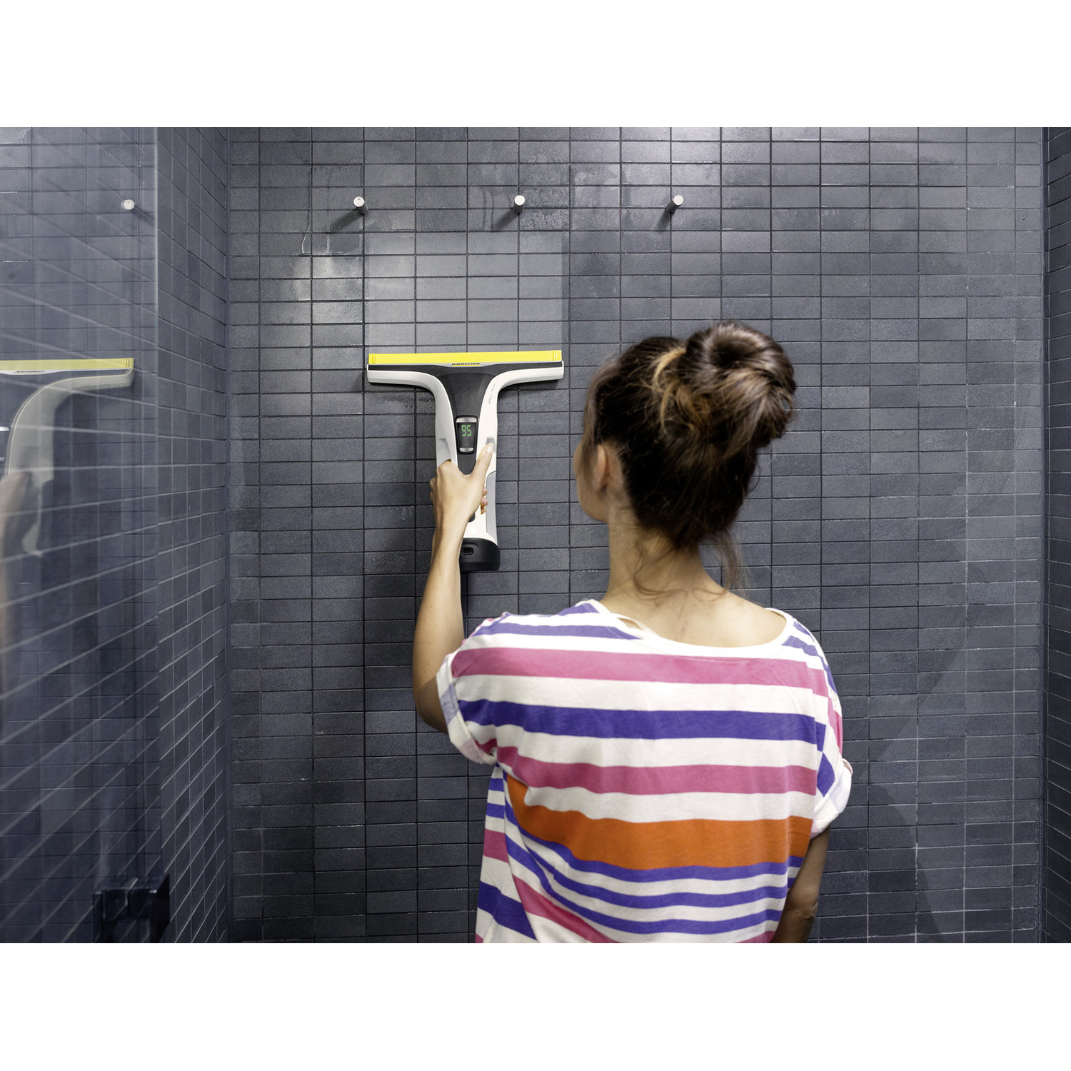 A woman cleaning a dark tiled shower wall with a yellow and white cleaning tool. She wears a striped shirt. The wall is wet.