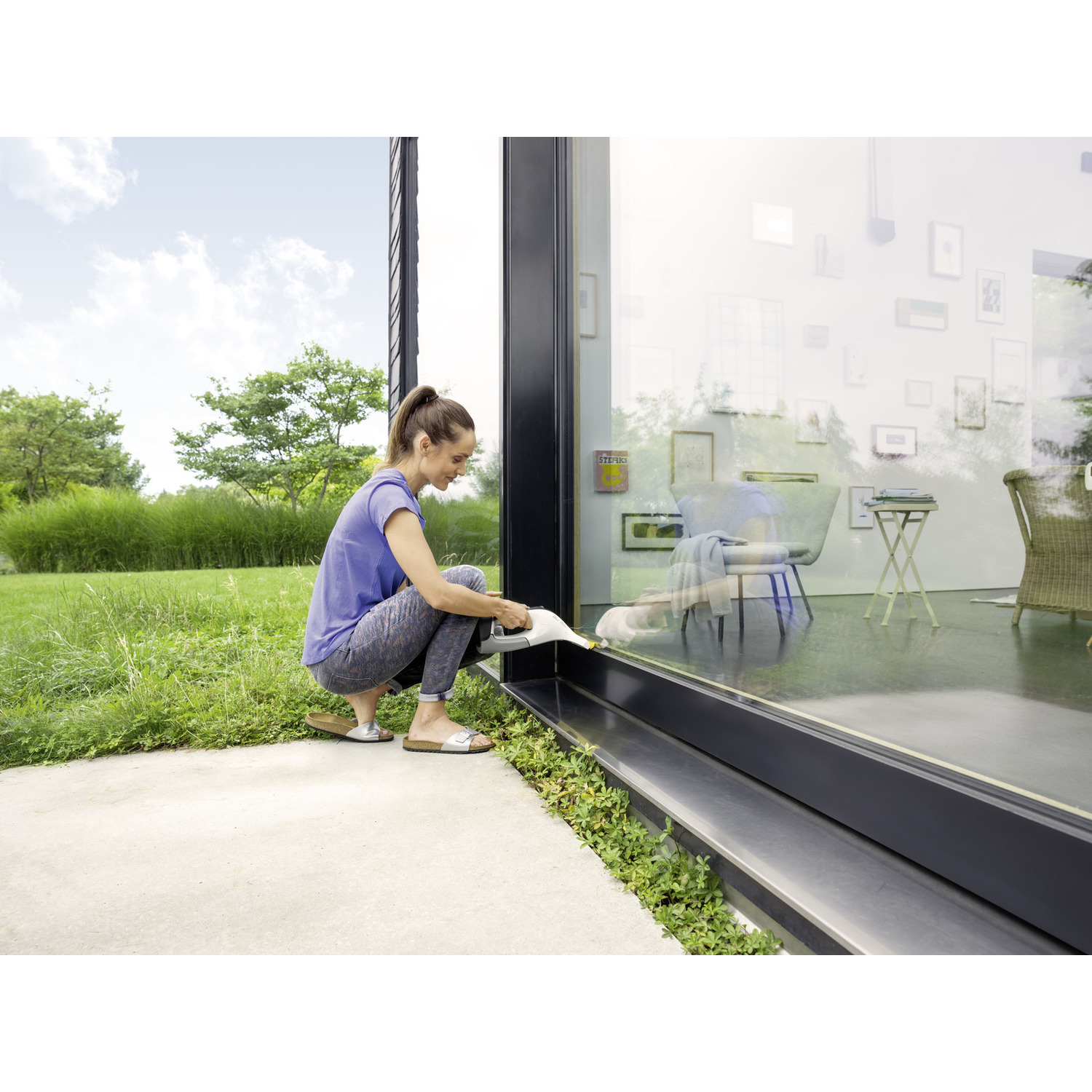 Woman cleaning a large window with a cleaning device. She is outside, grass and trees in background. Inside, chairs and art.