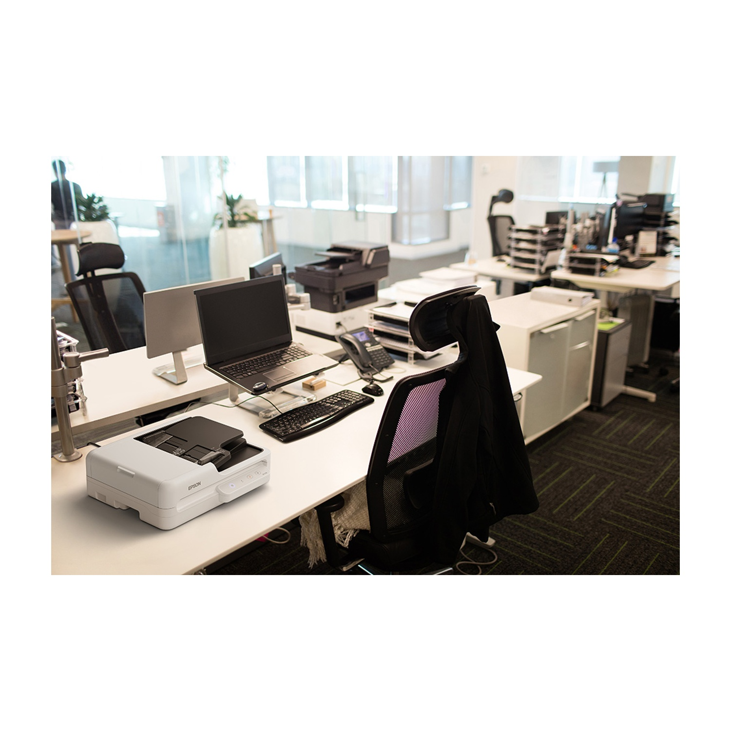 Un'area di lavoro in ufficio con scrivania bianca, laptop, stampante, telefono e sedia. Altre scrivanie e attrezzature per ufficio sullo sfondo.