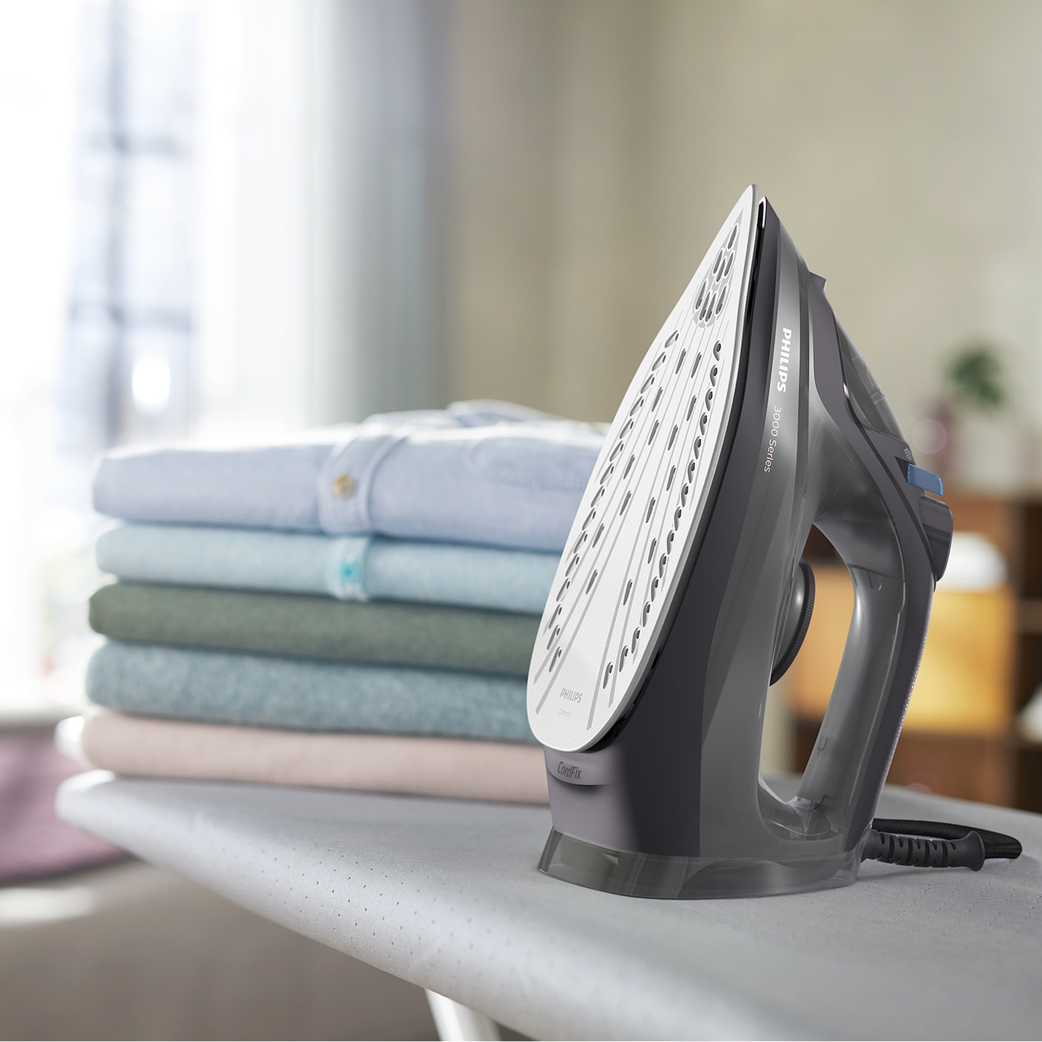A gray iron on an ironing board next to a stack of folded shirts in various pastel colors. The background is blurred.