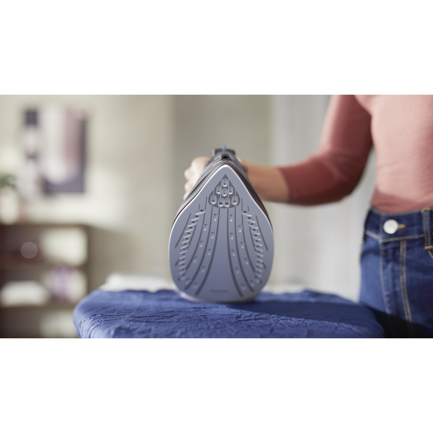 A person holds a gray iron over a blue ironing board. The person wears a red shirt and jeans.