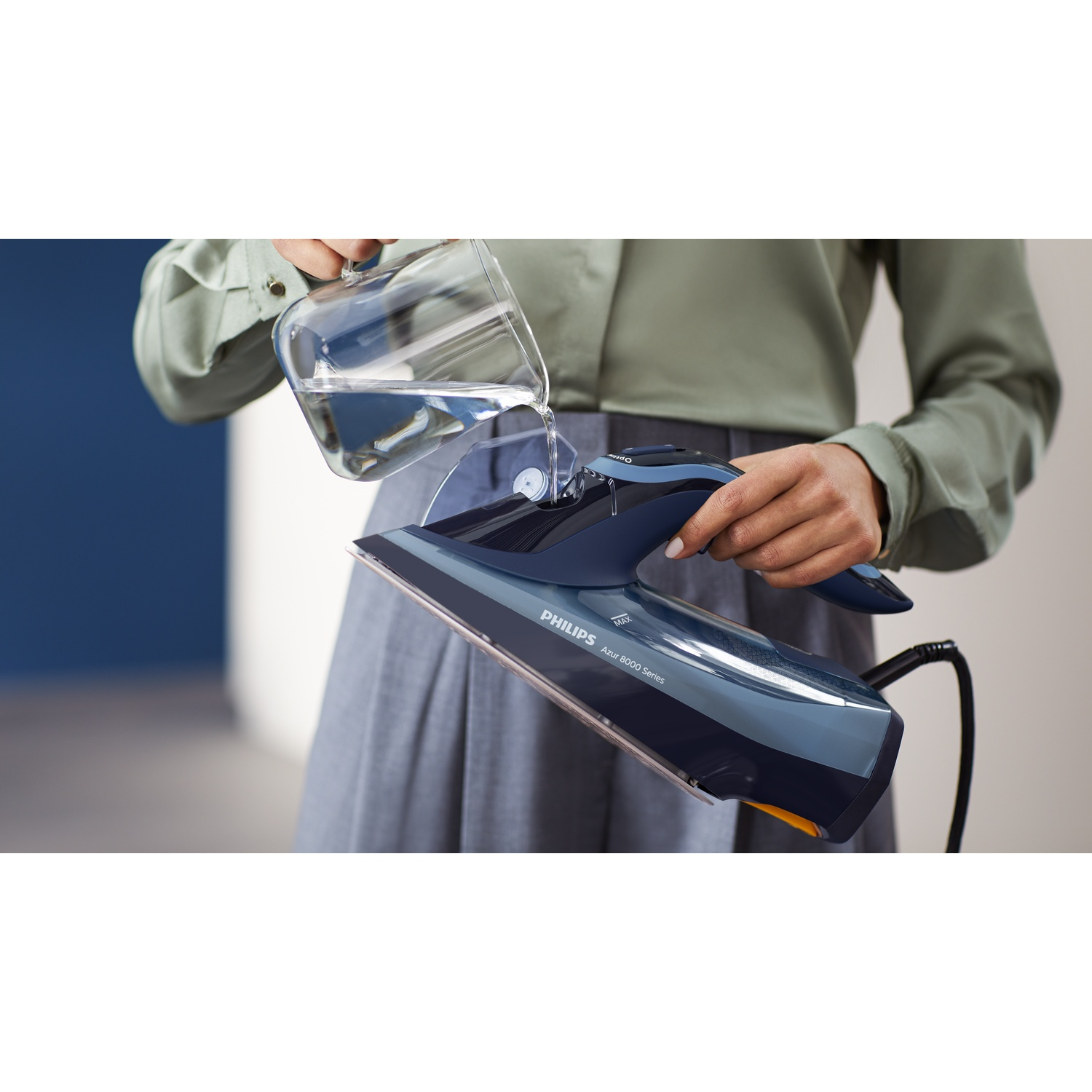 Une personne verse de l'eau dans un fer à repasser bleu et gris. La personne porte une chemise verte et un pantalon gris. L'arrière-plan est flou.