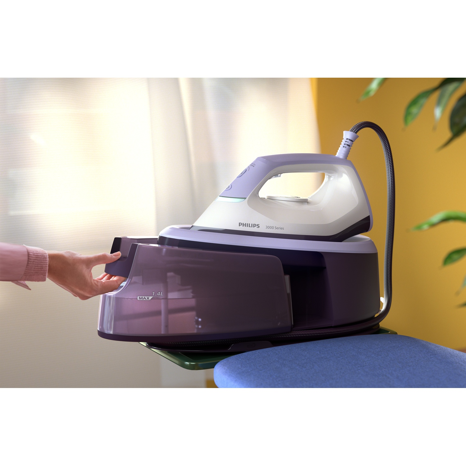 A person's hand is opening a purple steam iron's water tank. The iron sits on a green surface with a blue ironing board in front.
