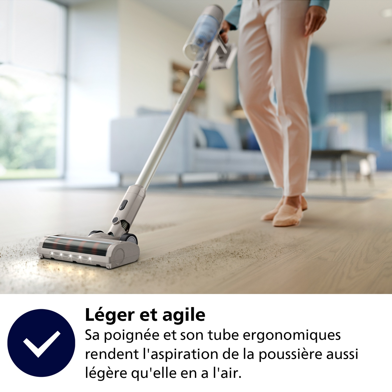 Une personne passe l'aspirateur sur un plancher en bois avec un aspirateur sans fil. Au fond, il y a un canapé et une fenêtre.