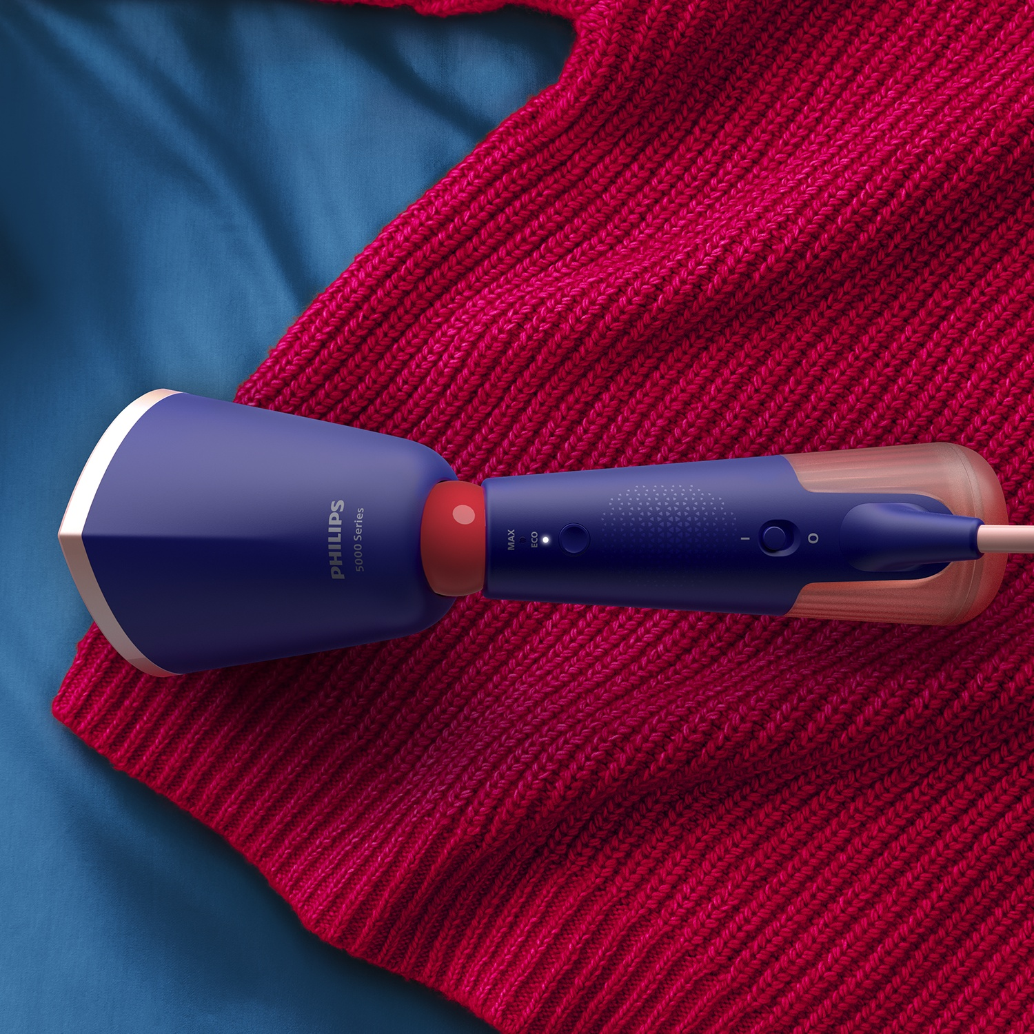 A blue Philips steamer on a red sweater, placed on a blue surface. The steamer has a red detail and a transparent handle.
