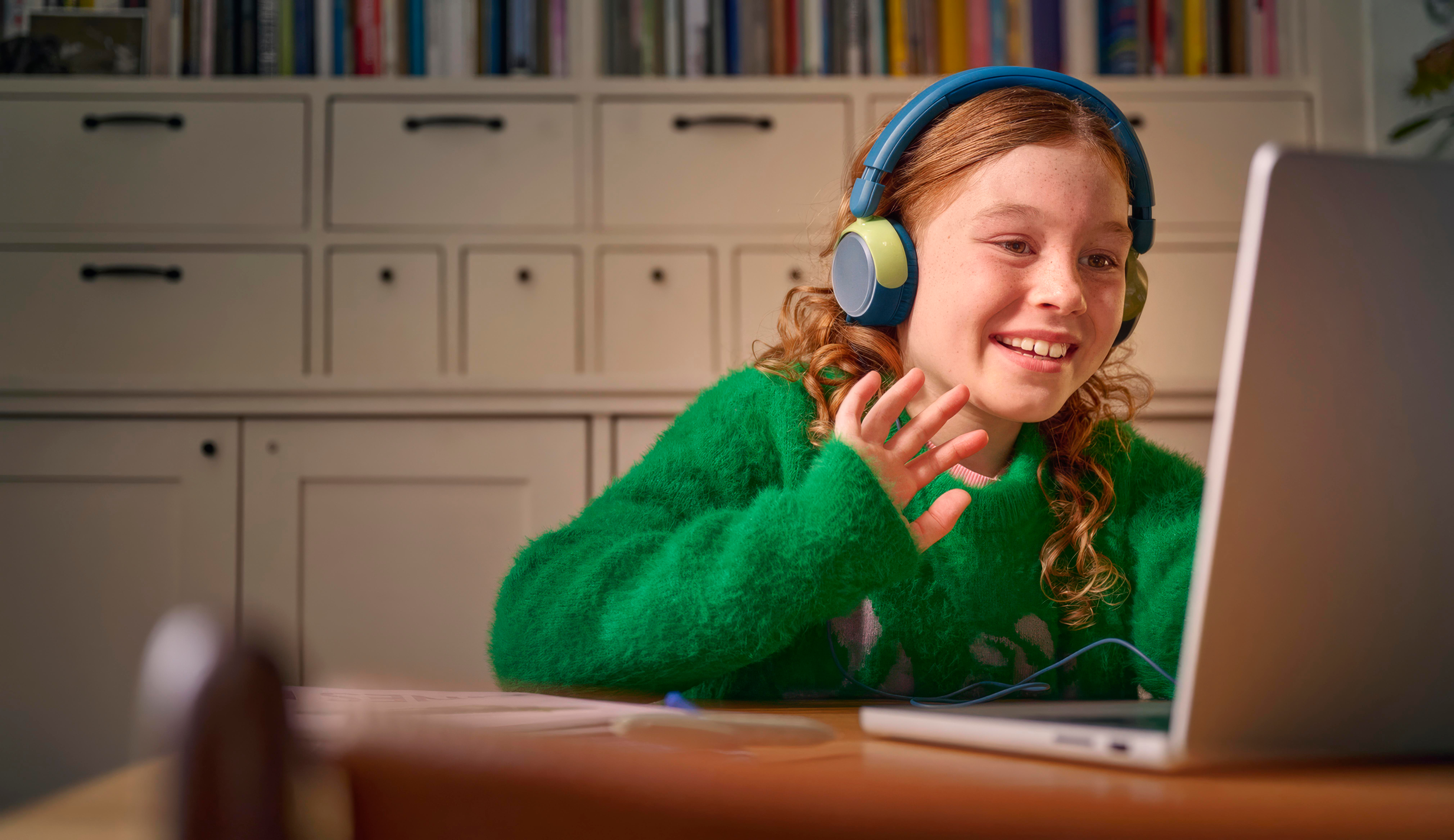 Ein Mädchen mit Kopfhörern lächelt auf einen Laptop-Bildschirm. Sie trägt einen grünen Pullover. Im Hintergrund sind ein Bücherregal und weiße Schränke zu sehen.