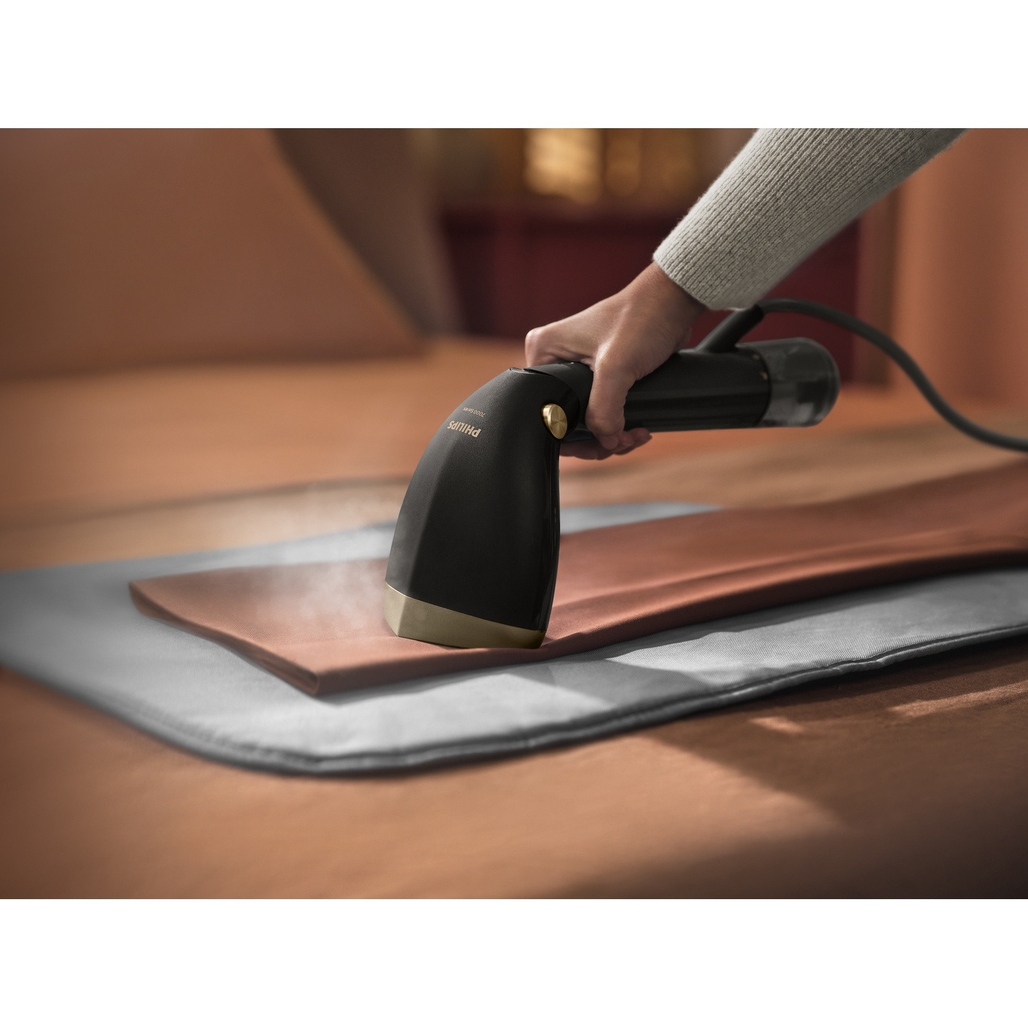 Une main tient un défroisseur noir et doré, qui défroisse un tissu marron placé sur une surface grise. L'arrière-plan est flou.