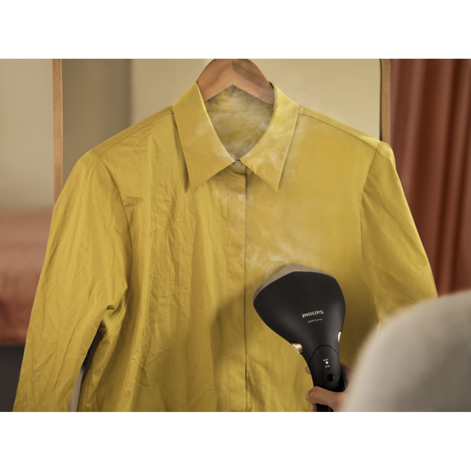 Un défroisseur Philips noir défroissant une chemise jaune suspendue à un cintre en bois. Vapeur visible. Une main tient le défroisseur.