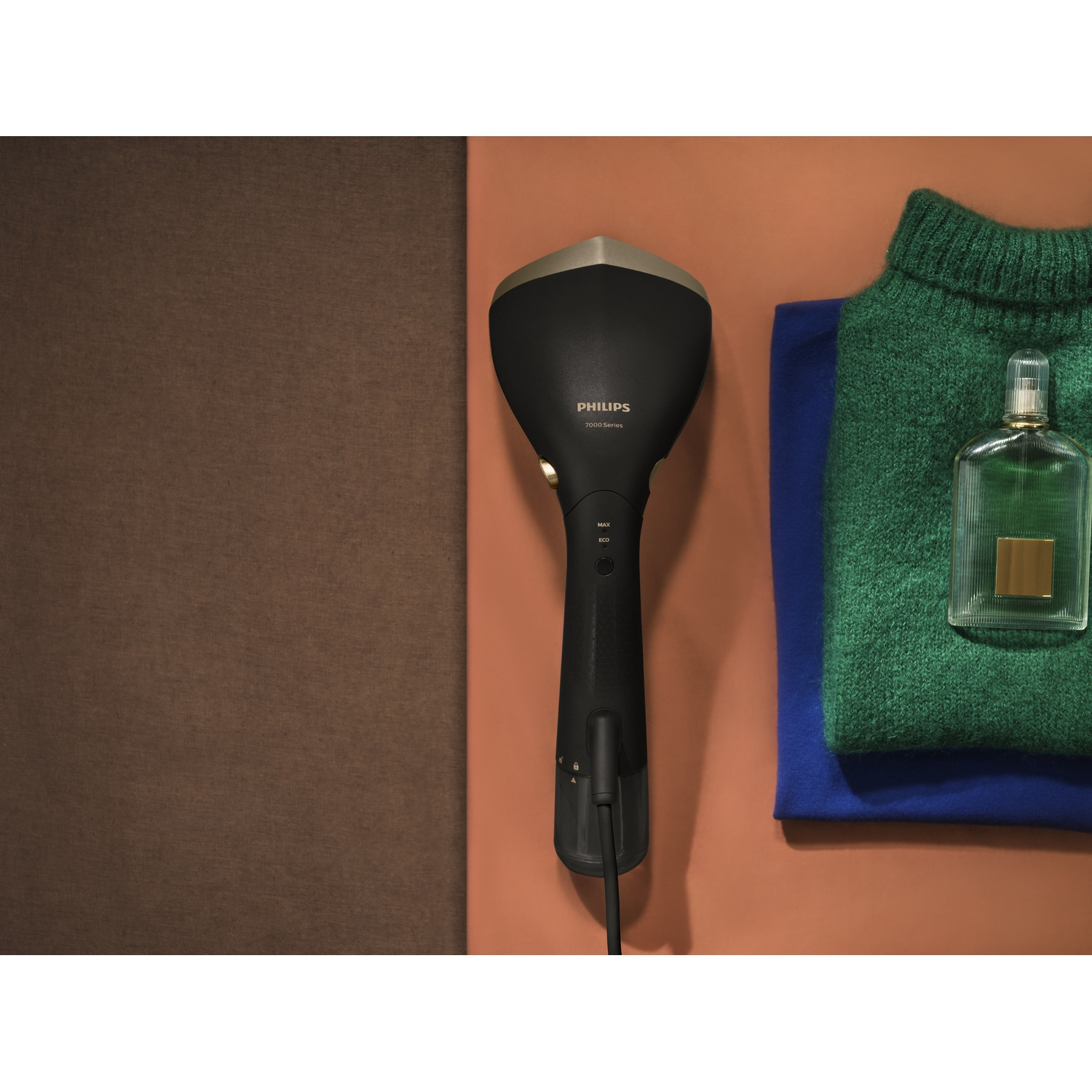 Un défroisseur Philips noir est à côté d'un pull vert, d'une chemise bleue et d'un flacon de parfum sur fond orange.