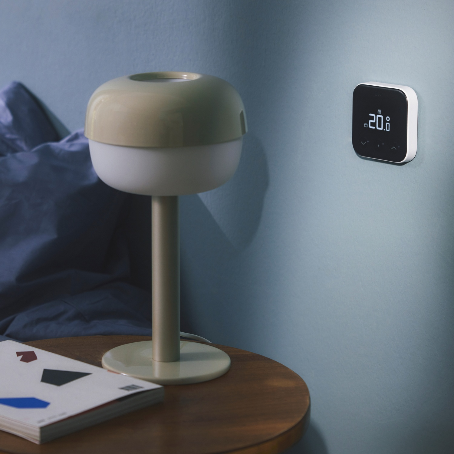 Un comodino con lampada e libro. Un termostato è montato sulla parete azzurra. Un letto sullo sfondo.