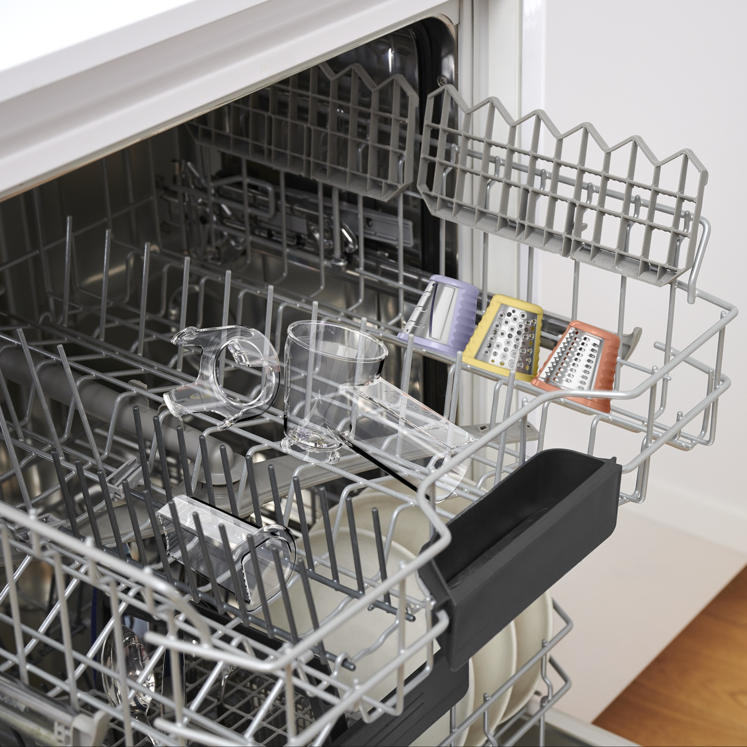 Dishwasher with clear glasses and colorful graters. Glasses are in the lower rack, graters in the upper rack. The machine is open.