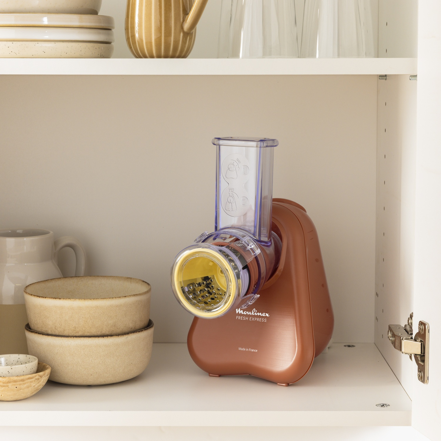 A copper Moulinex Fresh Express sits on a shelf. Bowls and plates are nearby. The scene is inside a white cabinet.
